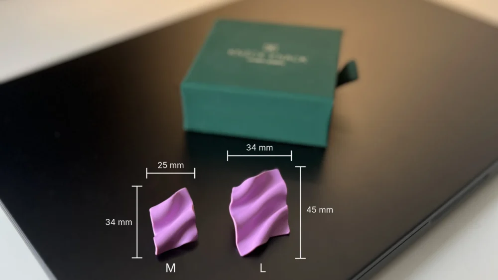 Two Soie earrings in M (34×25 mm) and L (45×34 mm) sizes on a dark surface with a jewelry box in the background