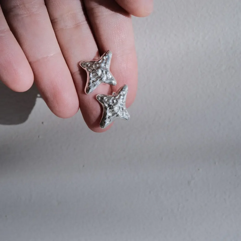 A close-up of a hand holding a pair of small, textured silver starfish-shaped earrings against a neutral gray background, highlighting their artisanal design and metallic finish.