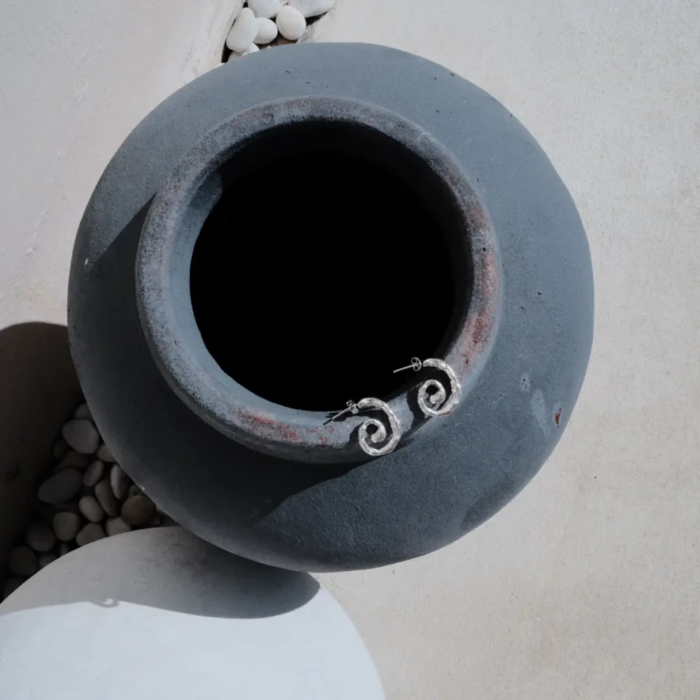 Handmade silver spiral earrings resting on the rim of a dark ceramic vase, surrounded by soft natural light and white pebbles