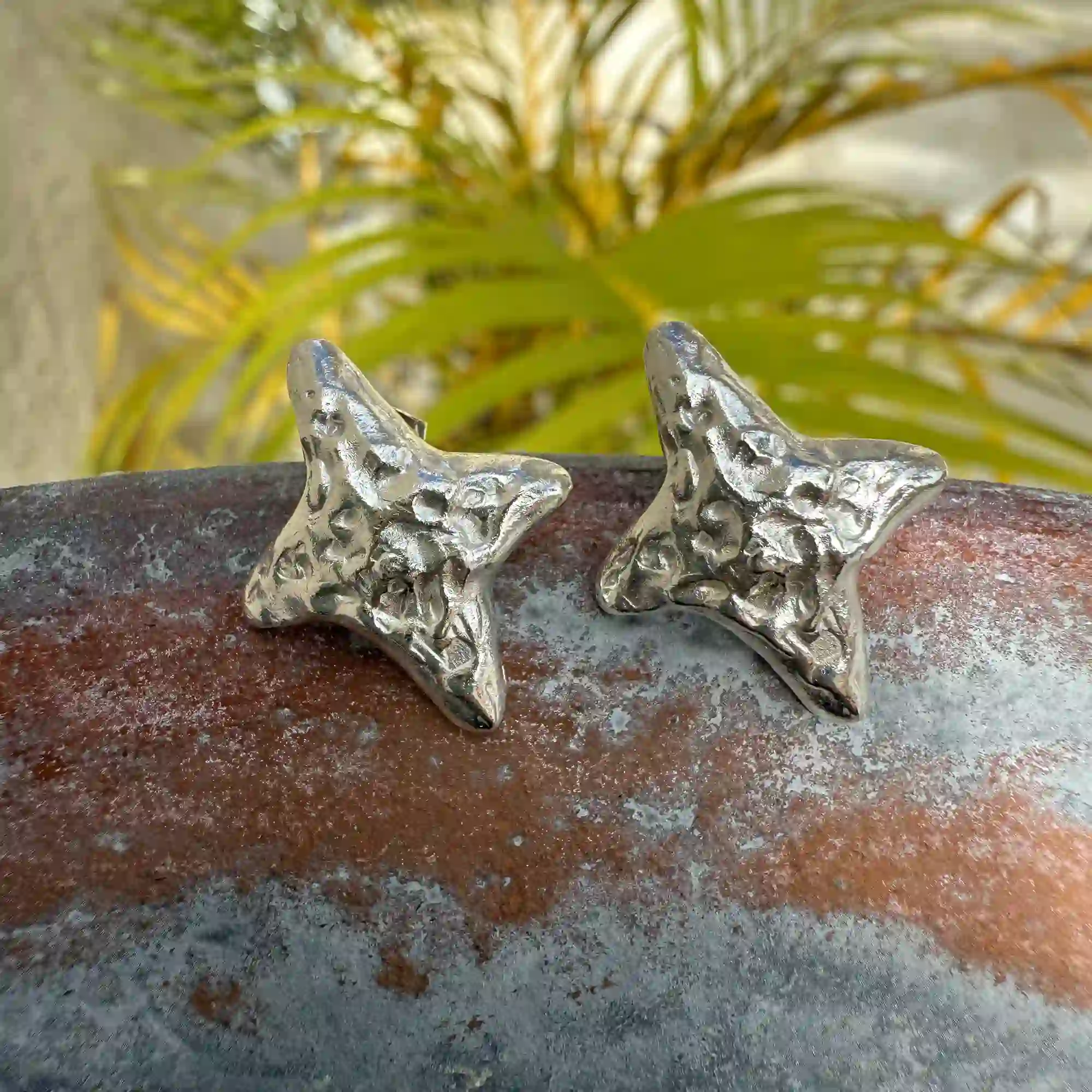 A pair of handcrafted, textured silver starfish-shaped earrings displayed outdoors on a rust-colored surface, with green tropical foliage blurred in the background