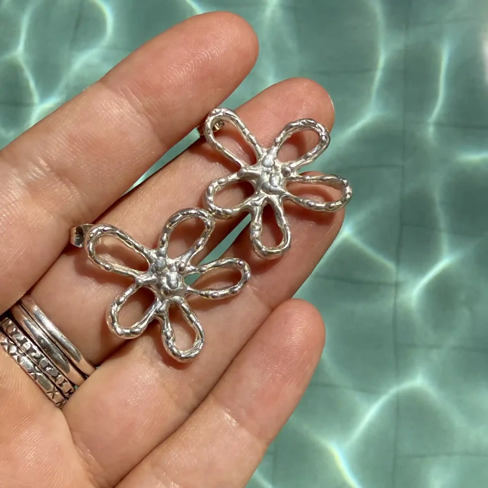 A pair of silver earrings shaped like abstract flowers with wavy, open petals, resting on a hand with sparkling water in the background.