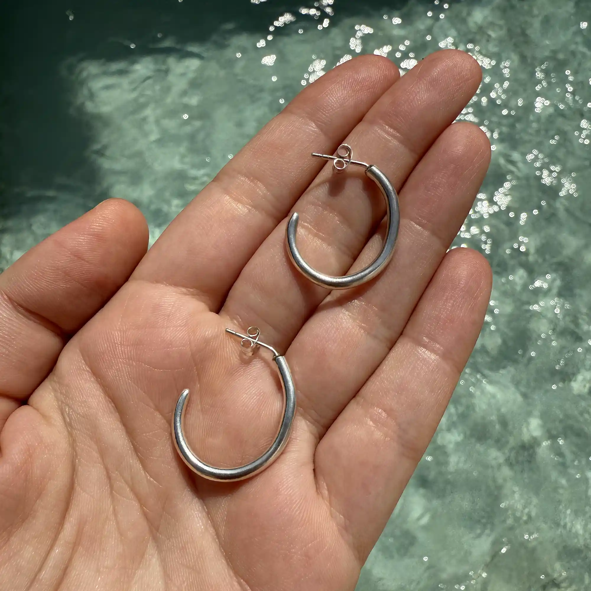 A pair of handmade silver hoop earrings with a slightly curved, minimalist design, displayed on an open palm with a sparkling water background.