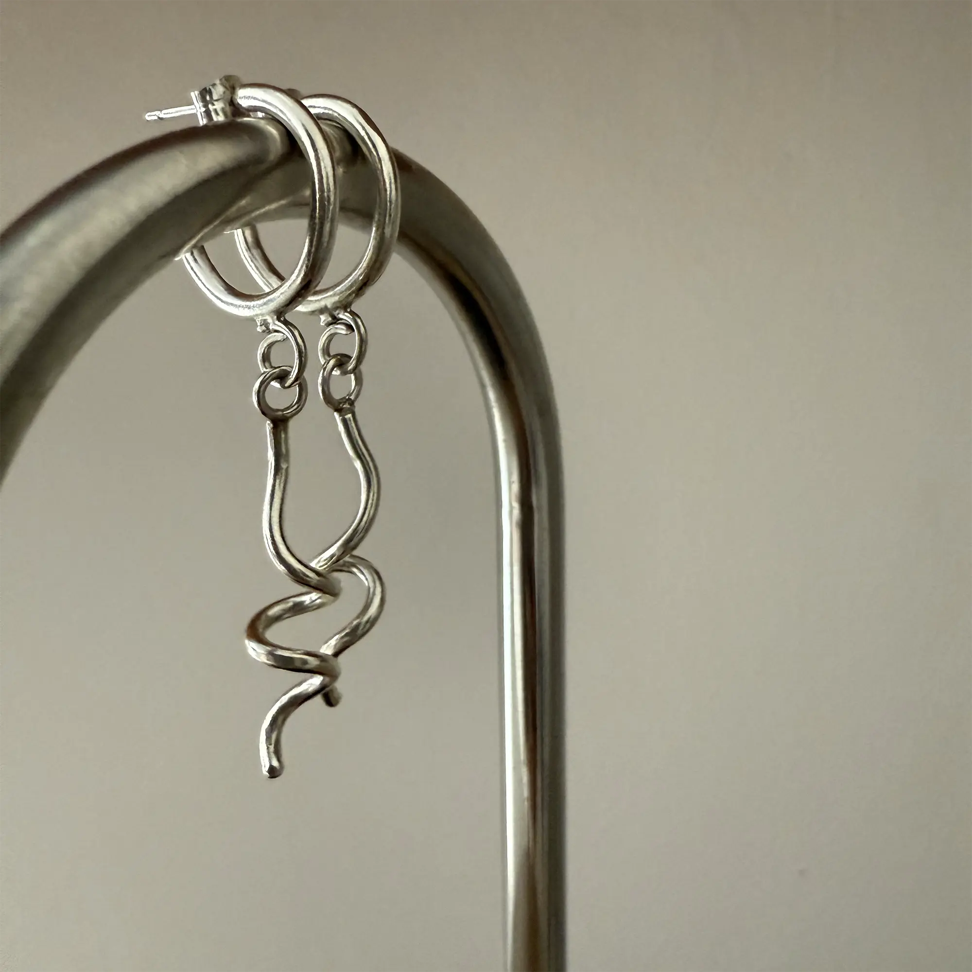 A pair of modern, abstract silver dangle earrings hanging from a curved metallic stand. The earrings feature twisted and wavy metal shapes with a minimalist, artistic design.