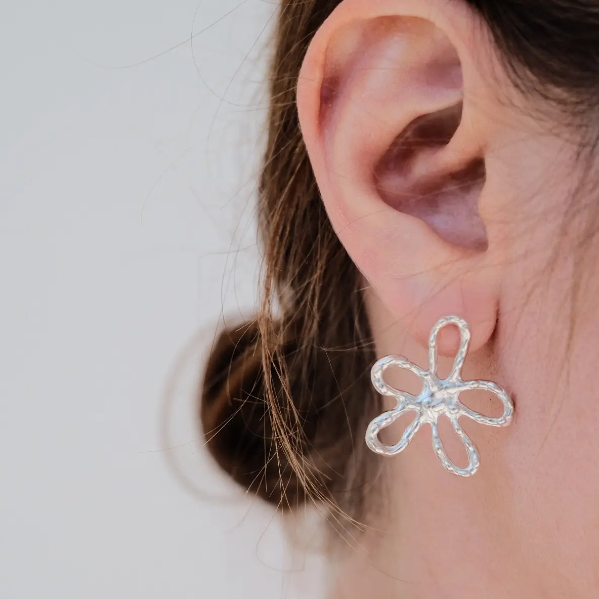 Close-up image of a woman's ear wearing a handmade silver earring shaped like an abstract flower with open petal loops and a hammered texture.